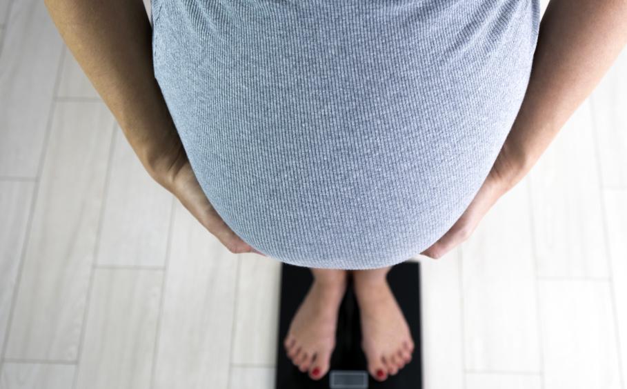 A woman's pregnant belly protrudes while she stands on a scale