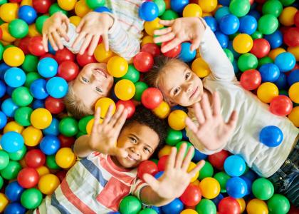 An overhead view of three young children laying in a pit of colorful balls with their hands extended into the air.