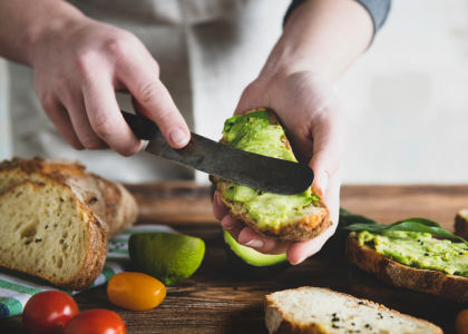 A man holds a piece of bread and uses a knife to spread pureed avocado on the bread. The table has a loaf of bread, tomatoes, and other slices of avocado and bread.