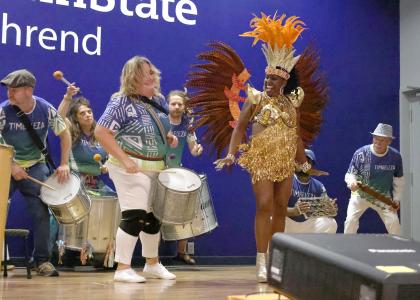 A costumed dancer jumps while on stage with several drummers.