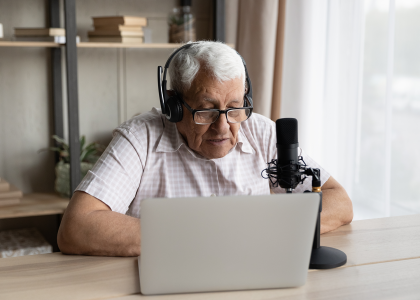 An older man sitting in front of a laptop wearing a headset and speaking into a microphone.