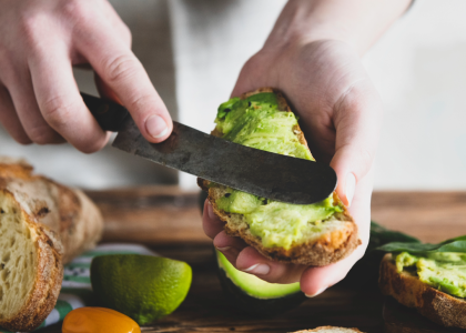 A person holds a piece of toast while spreading avocado on with a knife. The table has slices of bread, toast, and fruit. 