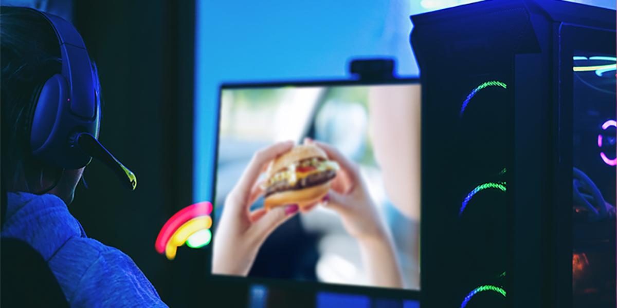 A person wearing a headset sits at a computer screen displaying two hands holding a cheeseburger.