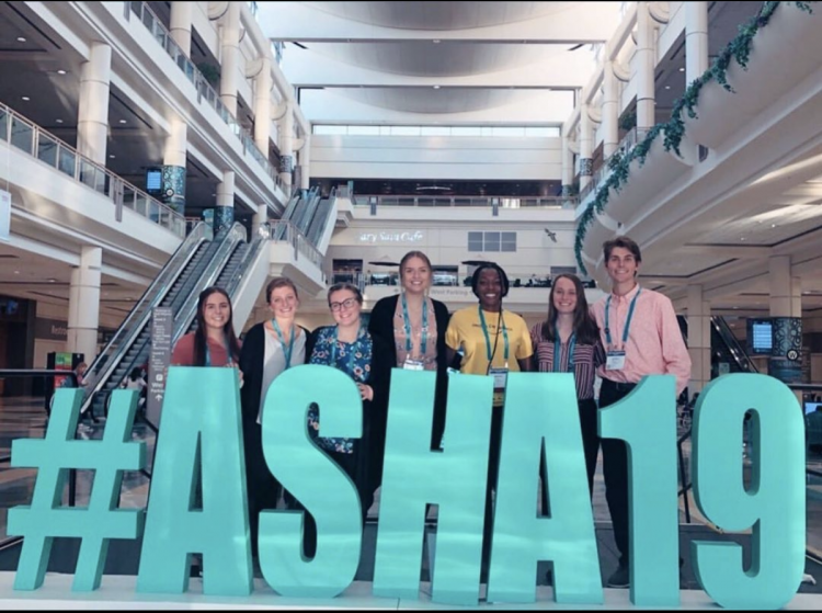 Group of students posed behind a large sign with ASHA conference hashtag