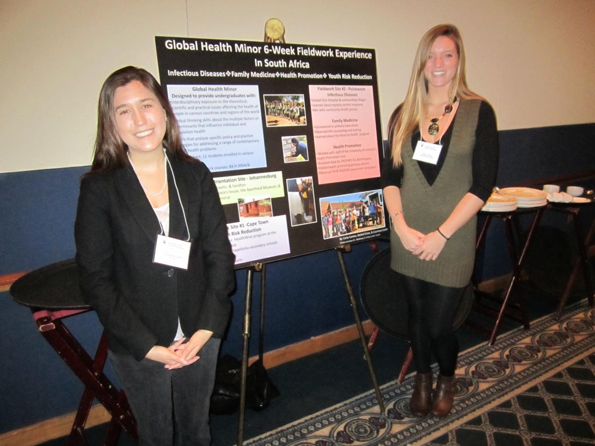 =Two female students standing in front of a presentation board titled "Global Health Mino 6-Week Fieldwork Experience in South Africa"